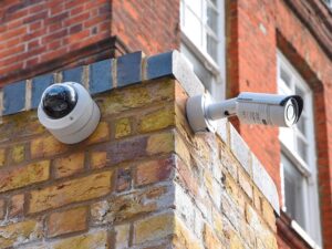Na imagem, duas câmeras de segurança realizam o monitoramento de um espaço externo.