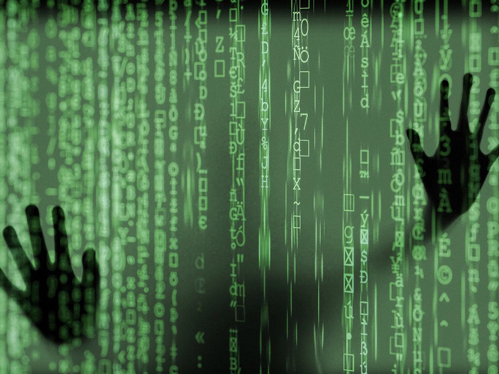 Imagens ilustrativa mostra diversos códigos em tela preta. Ao fundo, mãos humanas tocam a tela.
